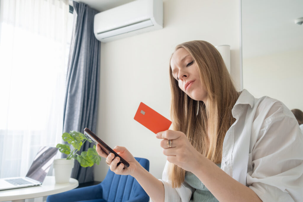 Woman checking phone and holding a card under a wall-mounted air conditioner, illustrating aircon using too much electricity and ways to reduce energy bills by Peakcool.